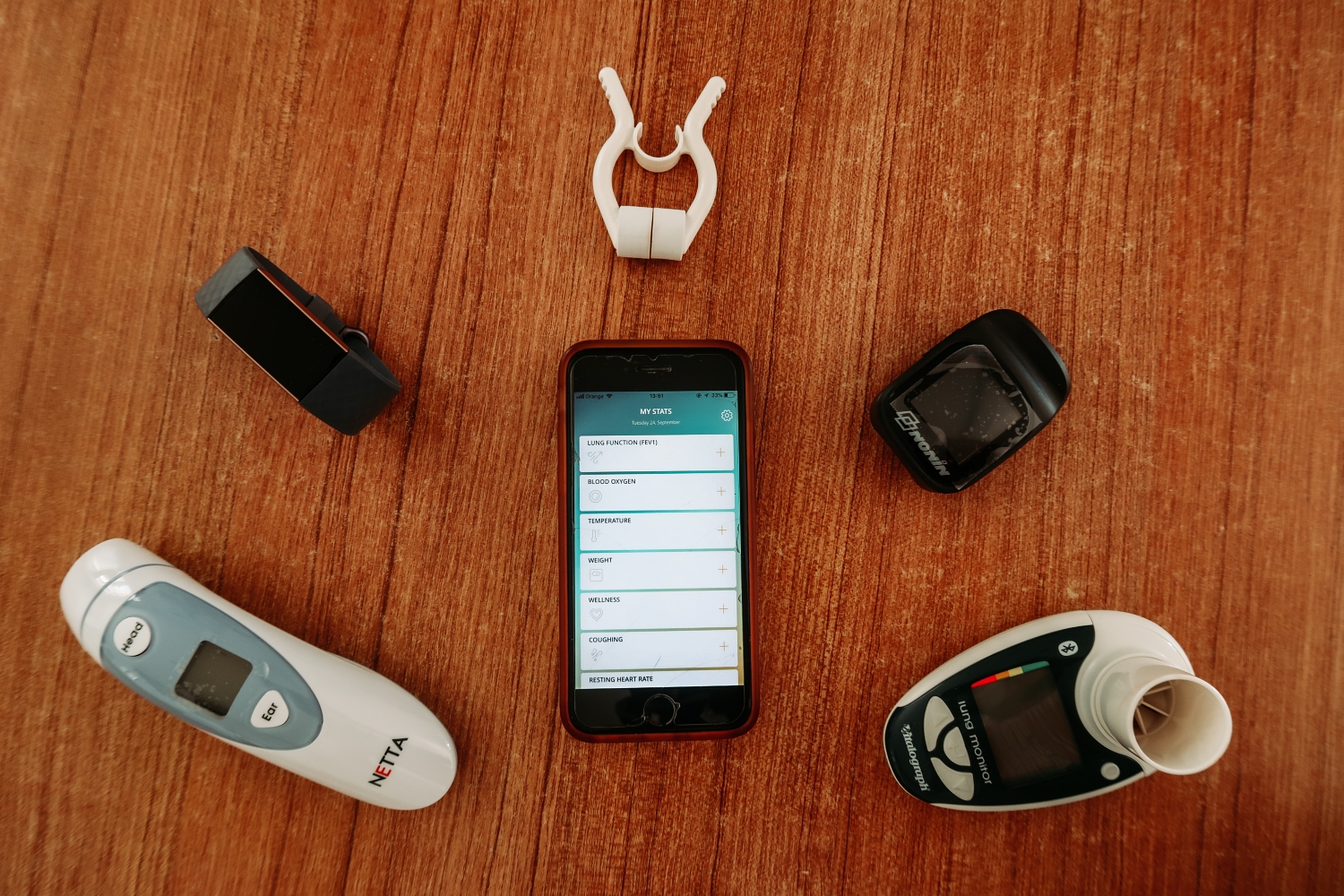 A smartphone on a brown desk, with a smart watch and thermometer.