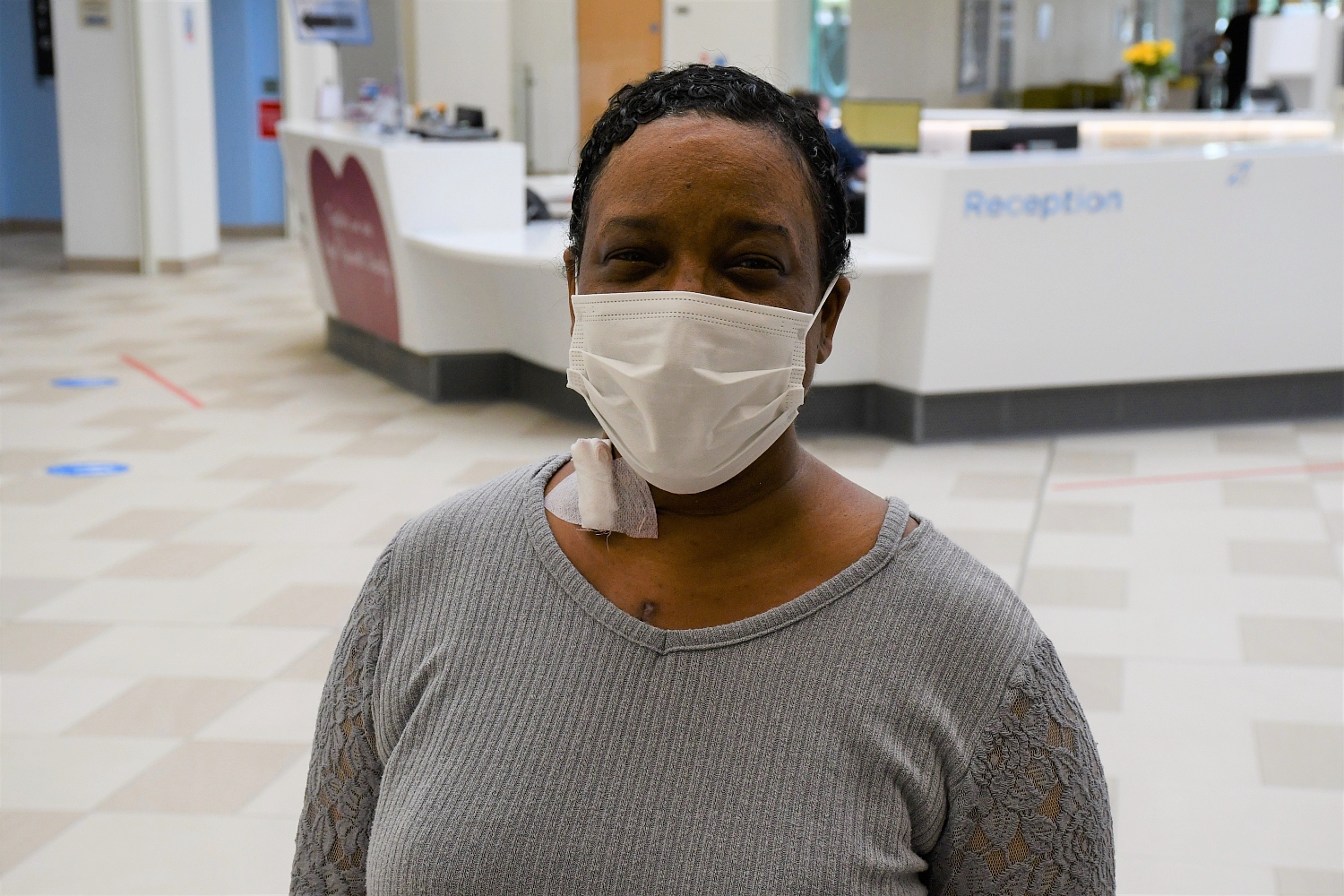 A woman with a mask smiles. In the background is a reception desk.
