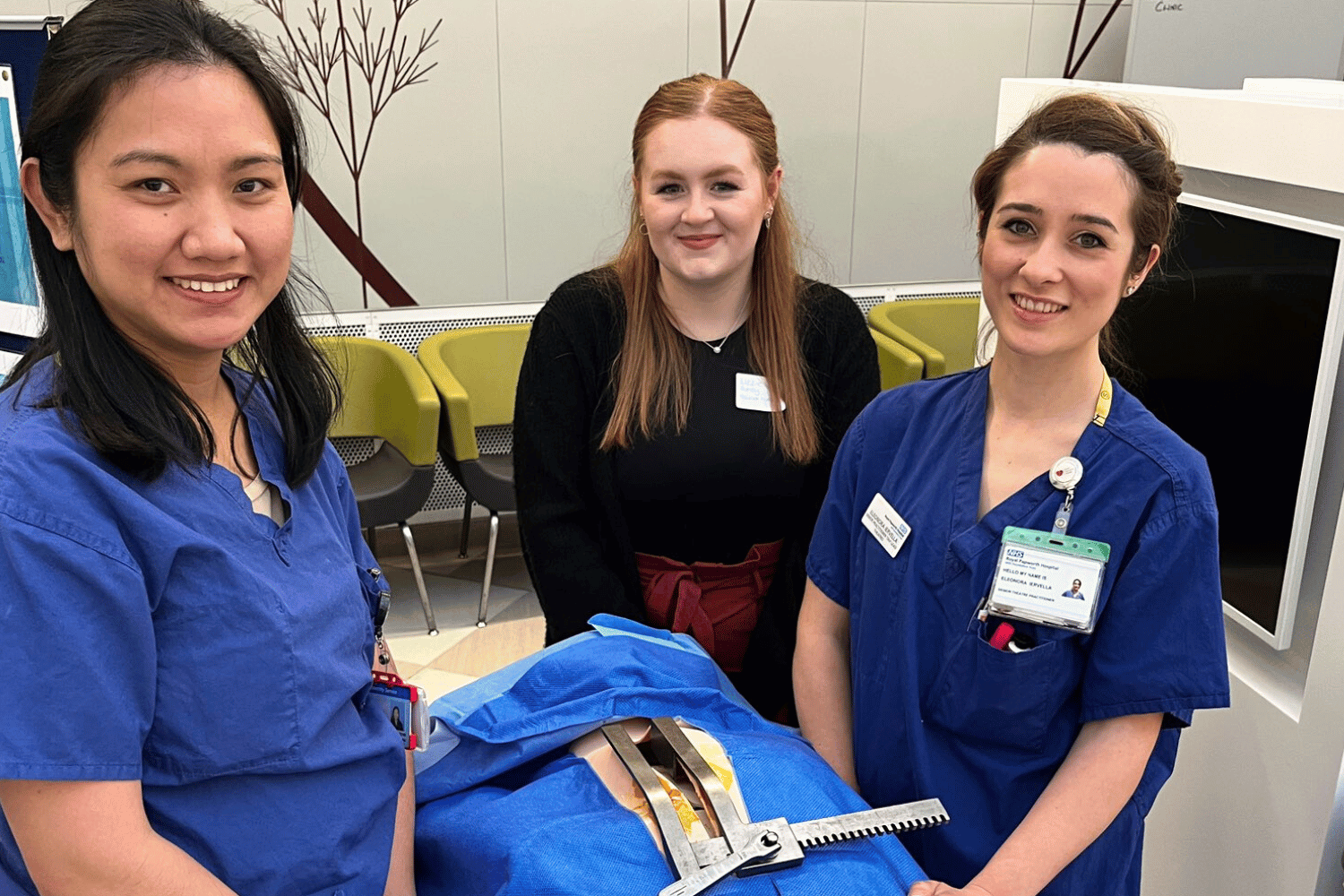 Three people standing inside, beside a fake body covered in blue surgical drapes.