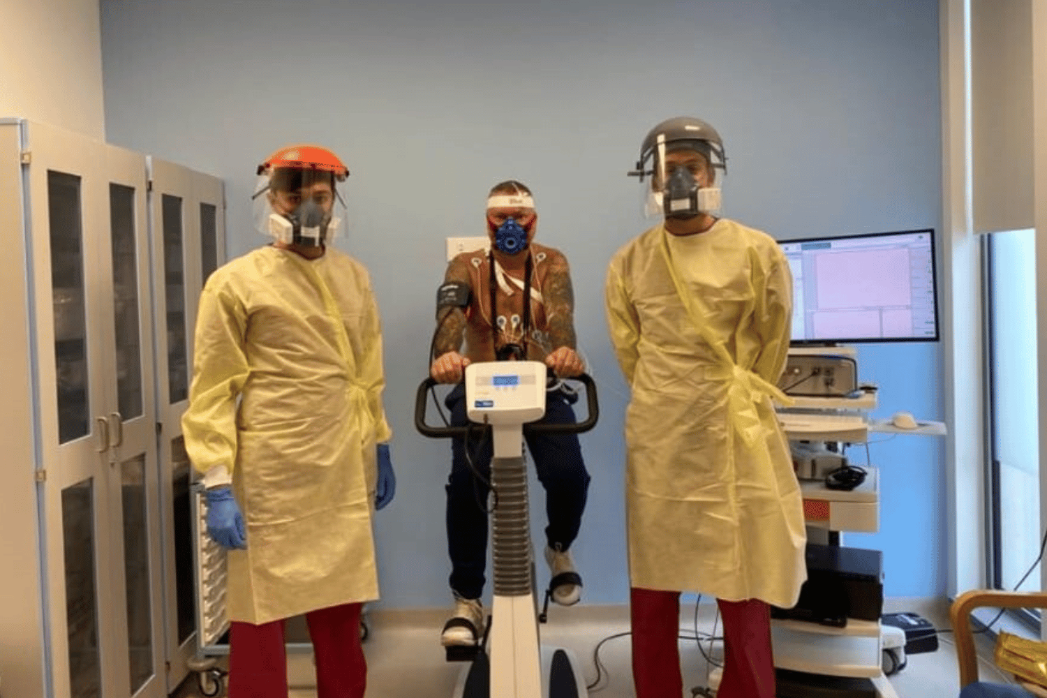 Two people in yellow plastic gowns, masks and visors with a man in the middle on an exercise bikes hooked up to wires connected to a computer.