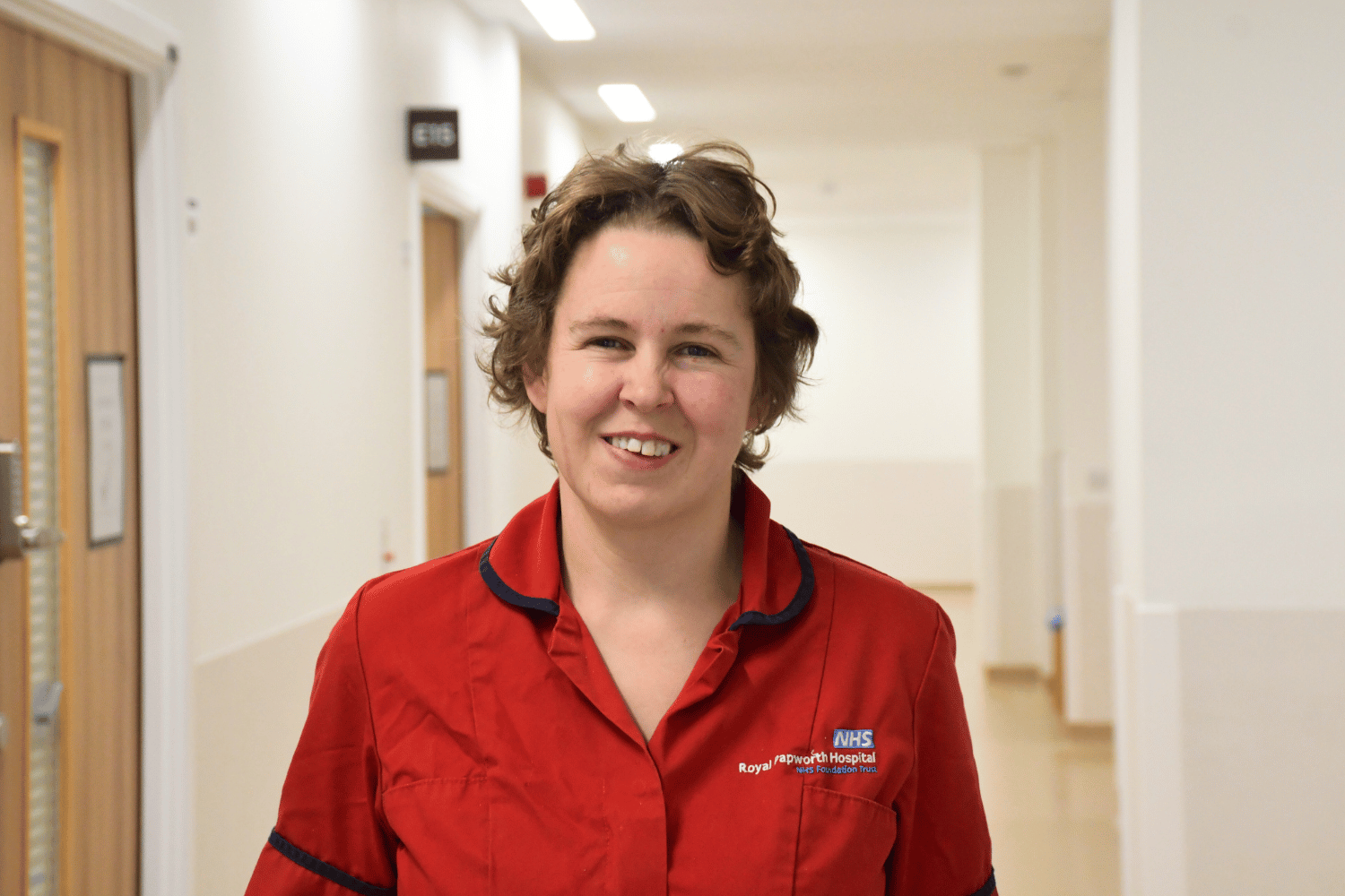 Lucy in a red top with navy blue trim smiling while standing in a hospital corridor.