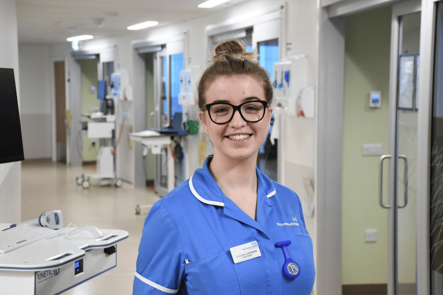 Claudia, wearing a blue nurse uniform and black spectacles, smiling in a hospital ward corridor.