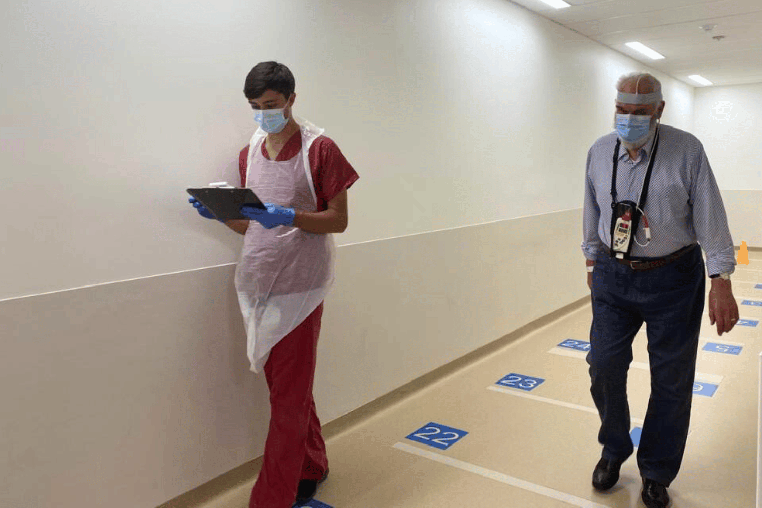 A person in red scrubs writes on a clipboard while a patient walks down a corridor lined with blue markers every metre