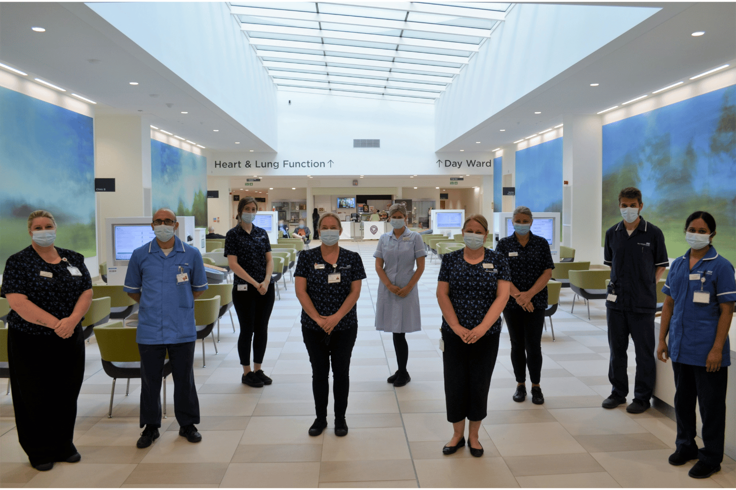 Nine healthcare staff in blue uniforms standing in a room with chairs and screens in the background.