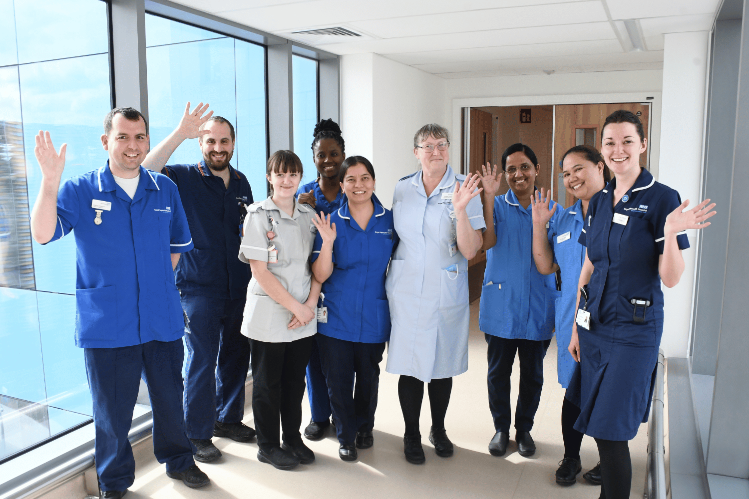 Nine nurses in different shades of blue smiling in a corridor. 