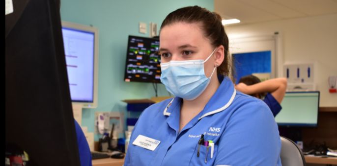 A nurse in a blue uniform and wearing a mask looking at a computer screen.