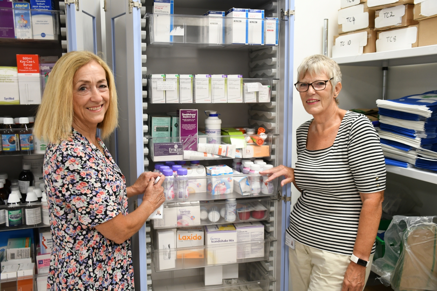 Two people standing either side of a cabinet full of boxes of medicines and drugs. 