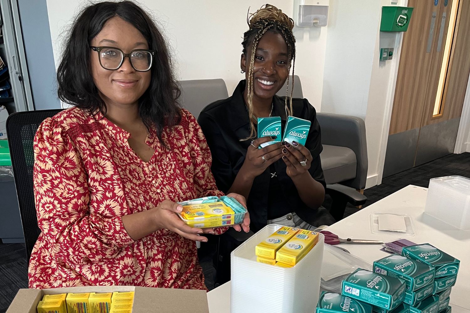 Two people smiling and holding up packets of sanitary products. 