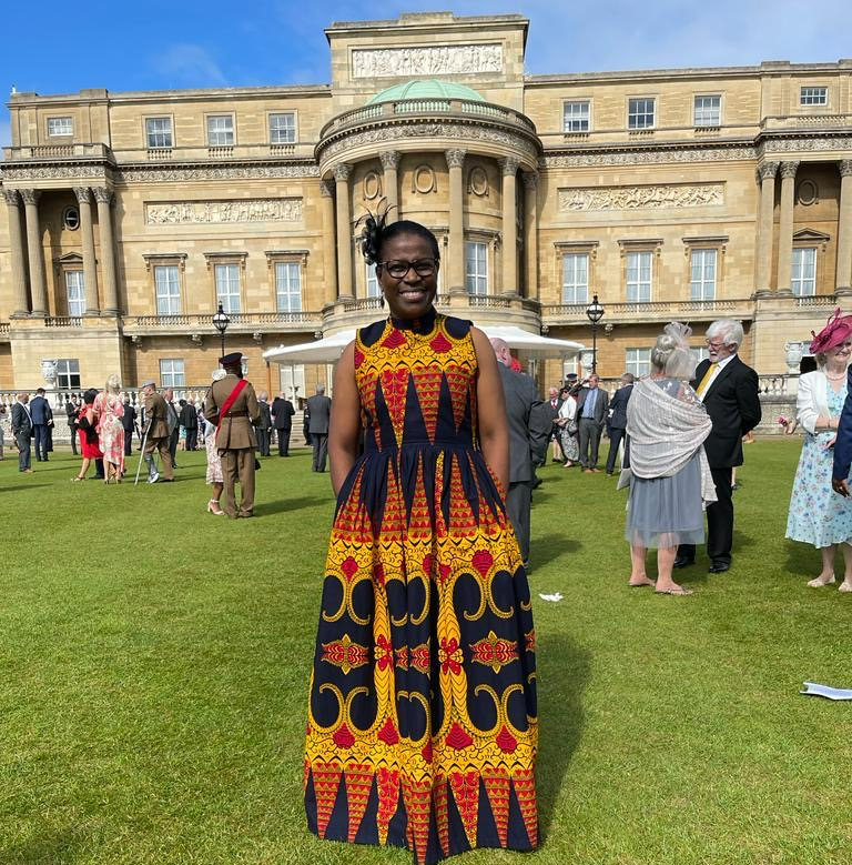 Judy in a navy blue, red and gold dress standing outside on grass, in front of a building and under a blue sky.