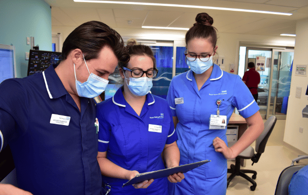 Three nurses talking while looking at a clipboard.
