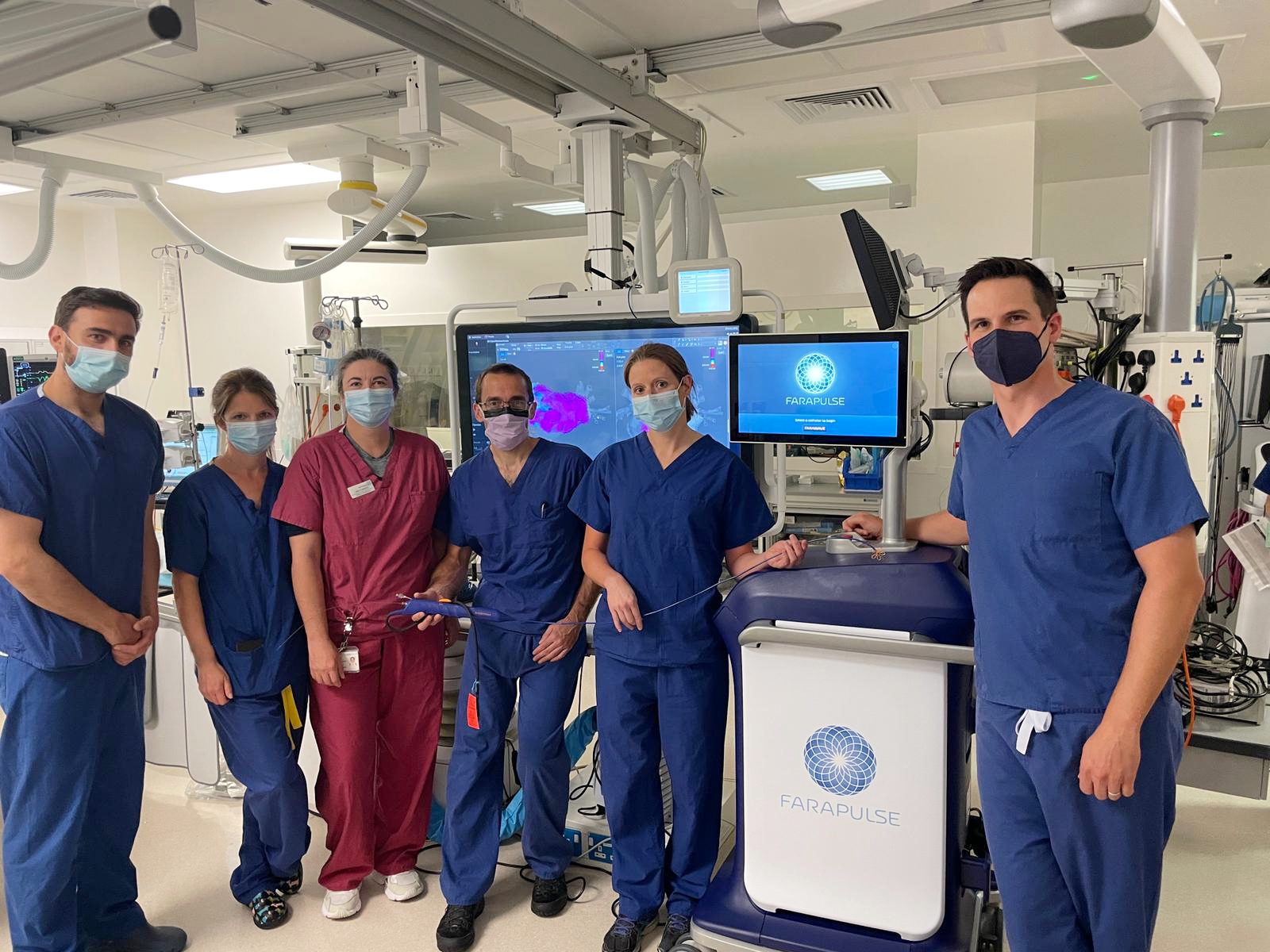 Six people standing in a line holding a long thin catheter. A screen in the background has an image of a heart.