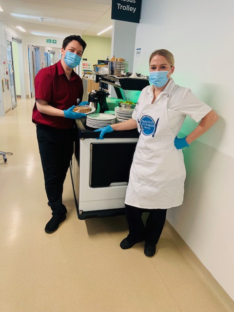 Two people standing in a hospital ward corridor next to a trolley full of food and plates.