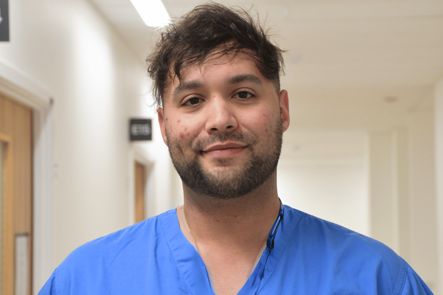 Will in blue scrubs standing in a hospital corridor smiling.