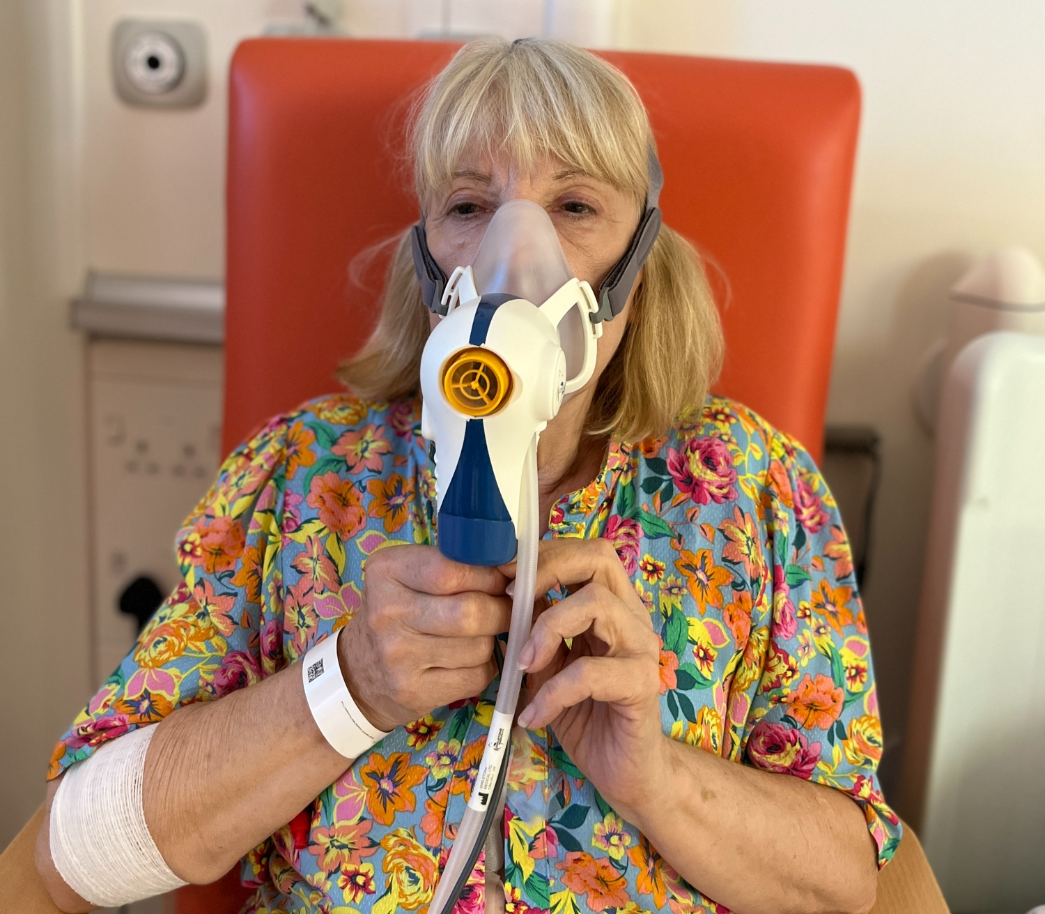 A woman in a colourful, flowery dress breathes into a blue and white plastic mask attached to wires and tubes.