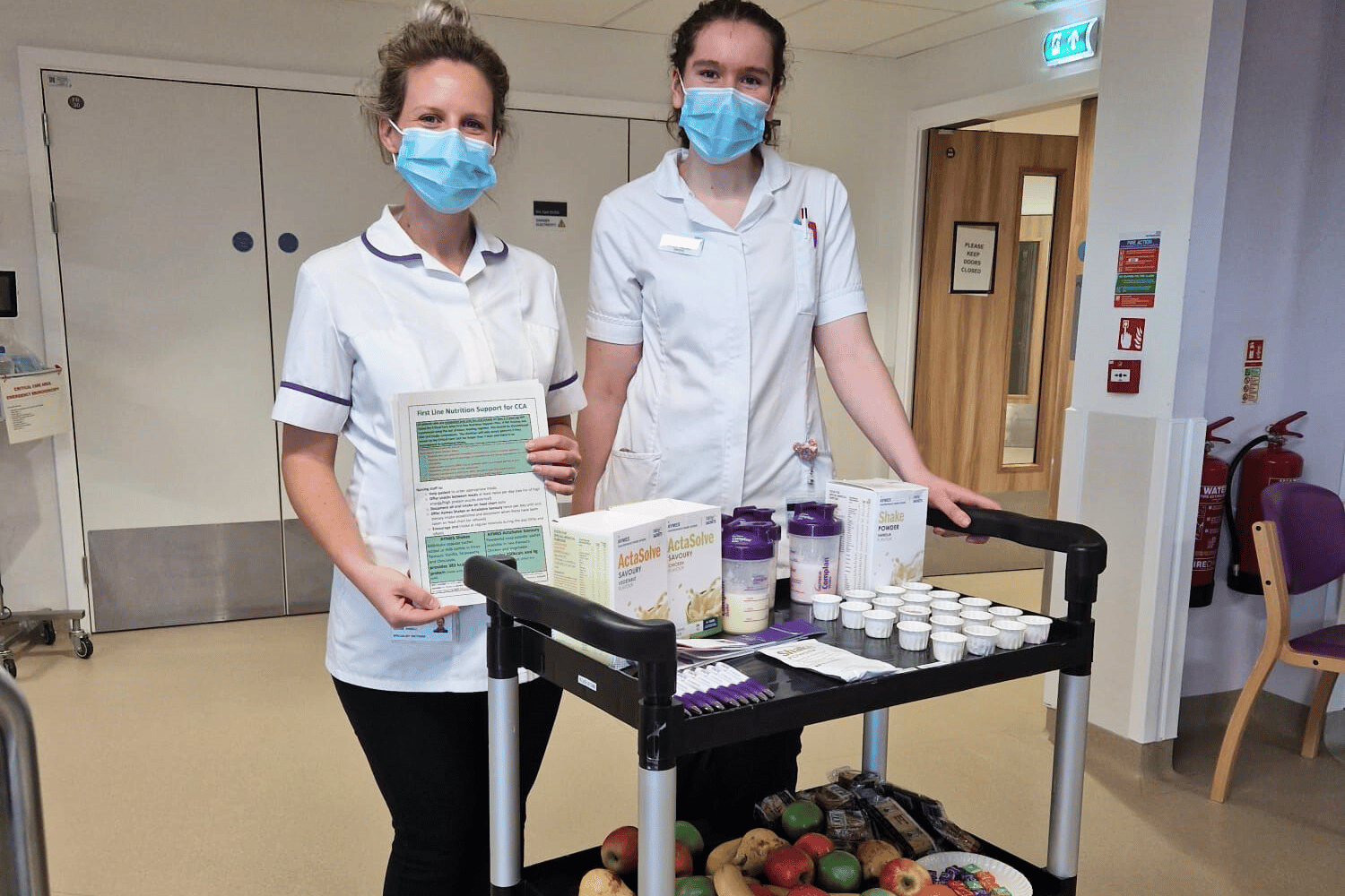 Two people in white uniforms with purple piping, wearing masks, standing with a trolley of fruit and drinks.