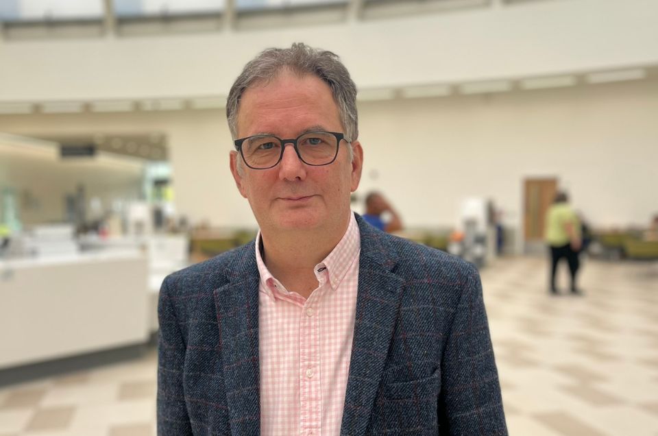 Alan, a heart transplant recipient, stands looking at the camera. He is wearing a pink chequered shirt and chequered jacket