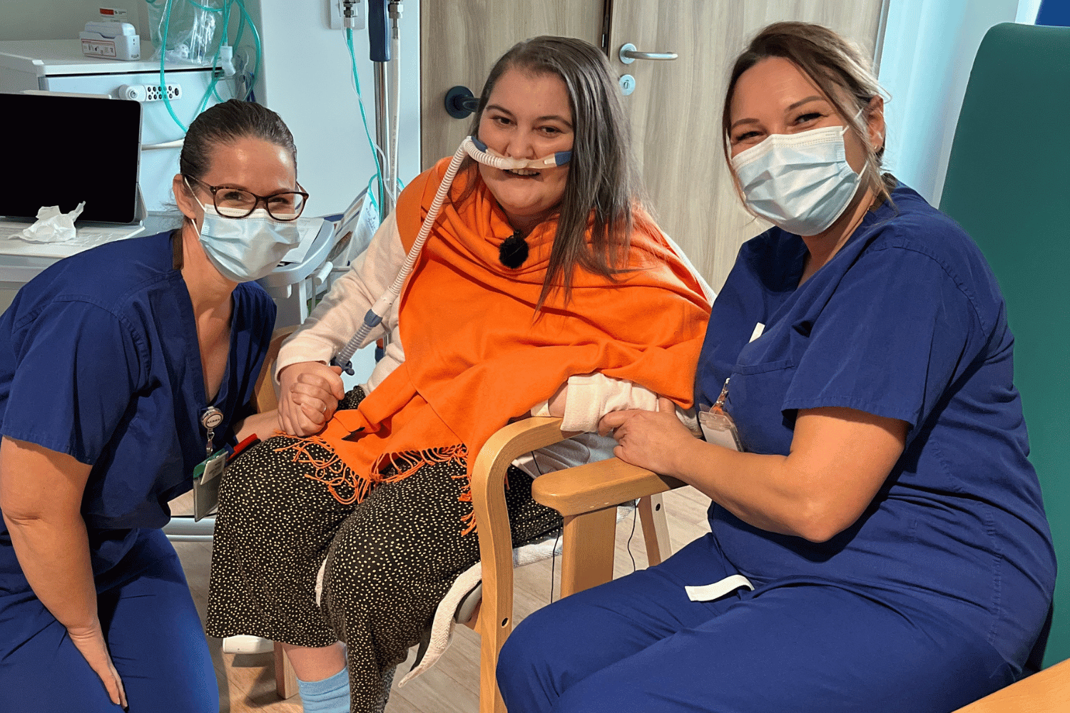 Two nurses in blue scrubs crouching either side of a patient in a chair.