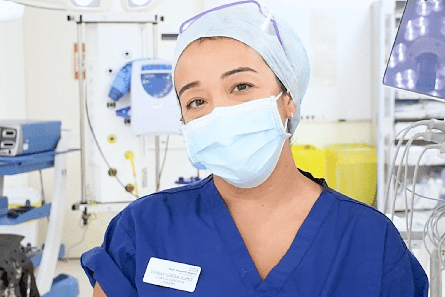 Tivisay in blue scrubs and wearing a face mask, with operating theatre equipment behind. 
