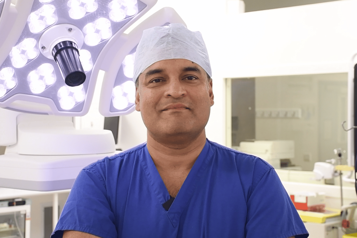 Shakil wearing blue scrubs and a theatre cap, smiling, with a theatre light behind him.